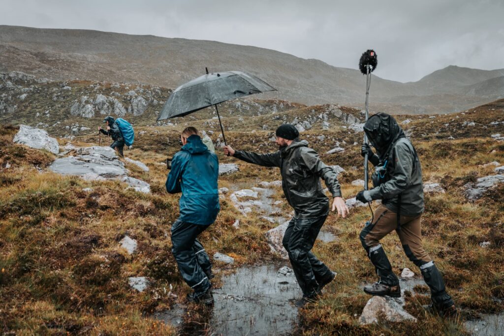 the north film crew stepping over a stream in the rain, while the director holds an umbrella over the camera