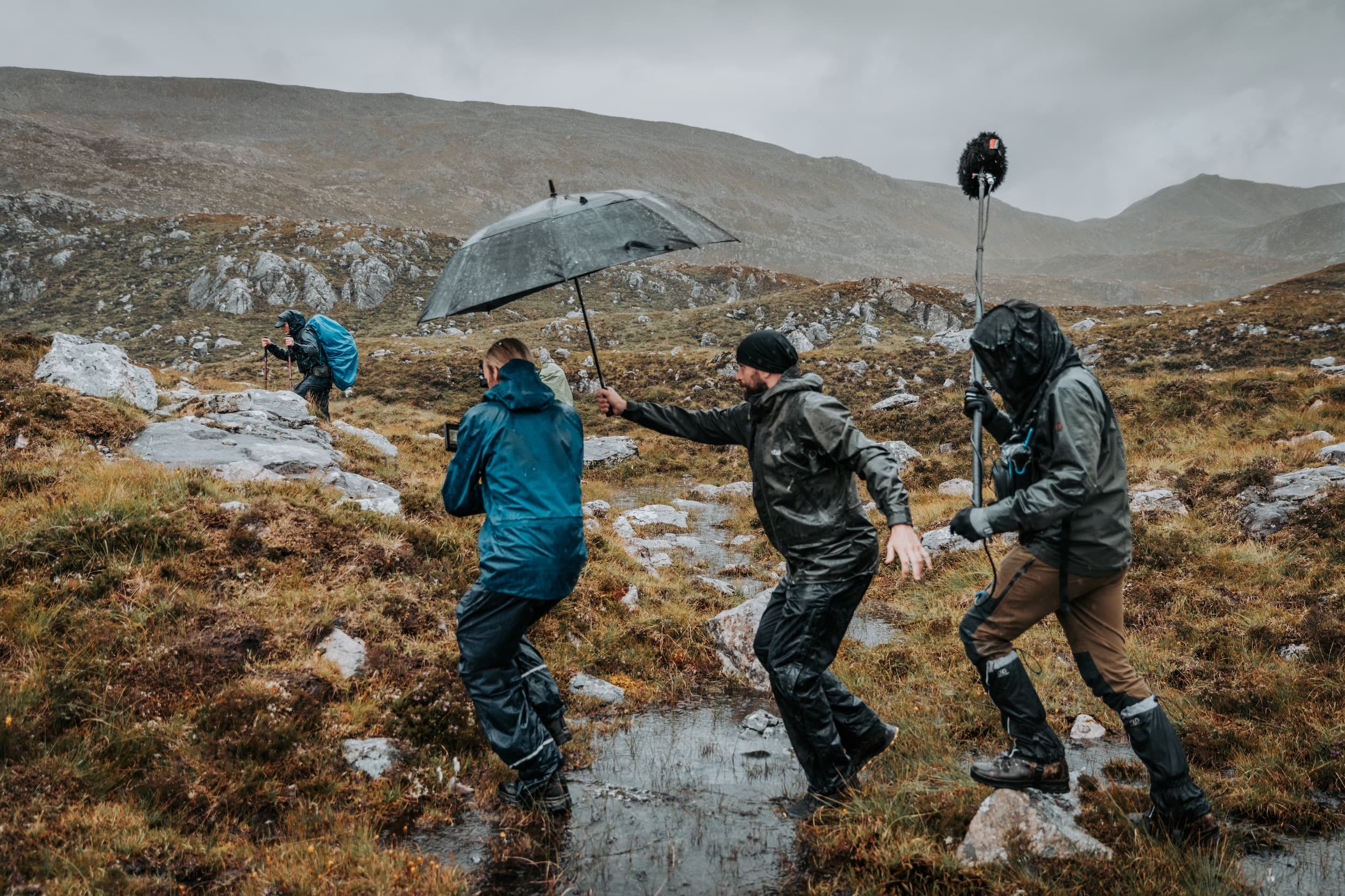 the north film crew stepping over a stream in the rain, while the director holds an umbrella over the camera